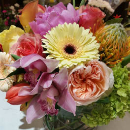 Short glass vase of mixed pink, yellow, peach, and green blooms including roses, gerbera daisy, orchids, and tulips.