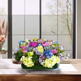 Low rectangular arrangement of pink, blue, and yellow flowers in a glass vase