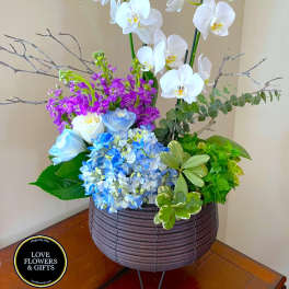Tall arrangement of white orchids, purple flowers, blue hydrangeas and white roses in a brown basket container.