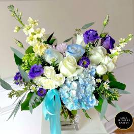 Mixed arrangement of white and blue roses, purple lisianthus, and blue hydrangea in a glass vase with aqua ribbon