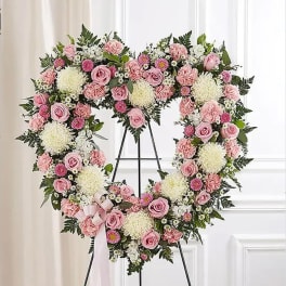 Heart-shaped standing wreath of pink roses, carnations, and white mums on a metal easel with a pink ribbon bow