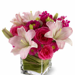 Compact pink lilies and roses arranged in a leaf-wrapped square glass vase