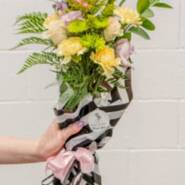 Hand-tied bouquet of pink lilies, yellow carnations, and green mums in striped wrap with a pink bow
