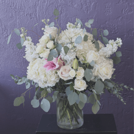White and blush bouquet of roses, hydrangeas, and lilies in a clear vase against a purple wall