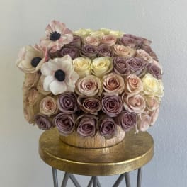 Round bouquet of blush, lavender, and cream roses with pale anemones in a gold box