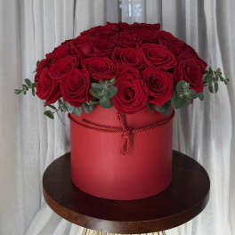 Red roses arranged in a round red box with eucalyptus accents