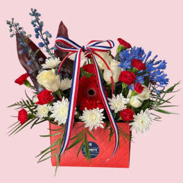 Red, white, and blue floral arrangement in a red gift box with ribbon