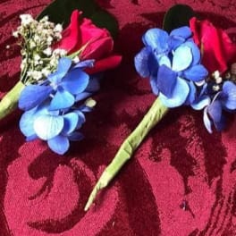 Two floral boutonnieres with blue flowers and red rose buds