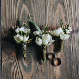 Three white floral boutonnieres on a wooden surface with two rings