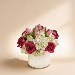 Low arrangement of pink roses, pink tulips, and white hydrangeas in a white cylinder vase
