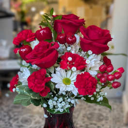 Tall red and white arrangement with red roses, red carnations, white daisies, and baby's breath in a red glass vase