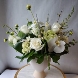 White arrangement of roses, hydrangea, and orchids in a white pedestal vase on a marble stand