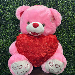 Pink teddy bear holding a red heart against a green hedge backdrop
