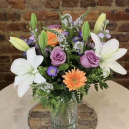 Mixed bouquet with white lilies, lavender roses, and orange gerbera daisies in a glass vase