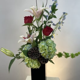 Tall floral arrangement with red roses, lilies, and blue flowers in a black vase