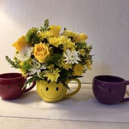 Yellow and white flower arrangement in a smiley-face mug, flanked by two colorful mugs