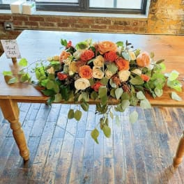 Long low bouquet of orange and cream roses with trailing greenery on a wooden table
