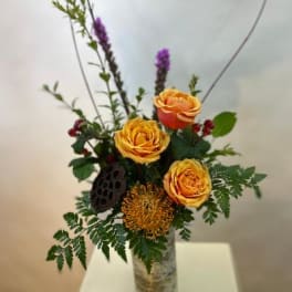 Orange roses in a tall glass vase with mixed greenery and purple accents