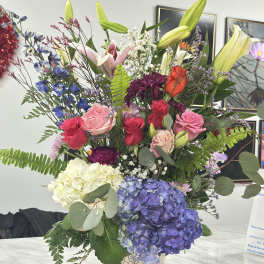 Mixed bouquet in a patterned paper cup with lilies, roses, hydrangeas, and other blooms
