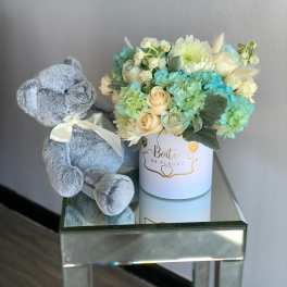 Blue teddy bear beside a pastel flower arrangement in a white box