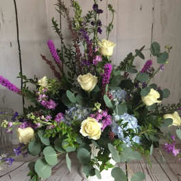 Arrangement of cream roses, blue hydrangeas, and purple spikes in a white vase