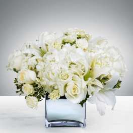 Low arrangement of white roses, hydrangeas, and lilies in a clear glass cube vase