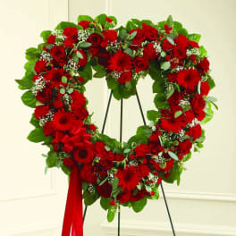Heart-shaped standing wreath of red roses and other red flowers with a red ribbon.