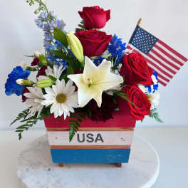 Red roses and white lilies in a patriotic wooden container with a small U.S. flag.