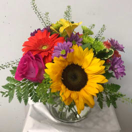 Mixed bouquet with sunflowers, roses, gerbera daisies, and purple blooms in a glass vase