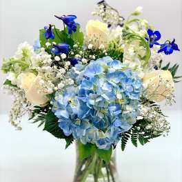 Vase arrangement with blue hydrangeas, cream roses, white blooms, and bright blue flowers