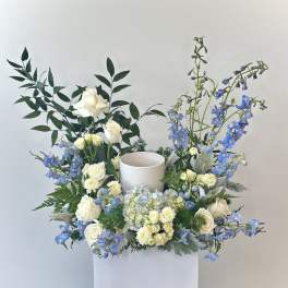 White roses and blue flowers arranged around a candle holder in a white box.