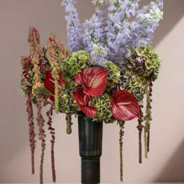 Tall arrangement with red anthuriums, green hydrangeas, and pale lavender flowers in a dark vase.