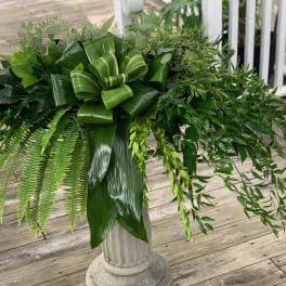 Green foliage arrangement in a pedestal vase with a large bow