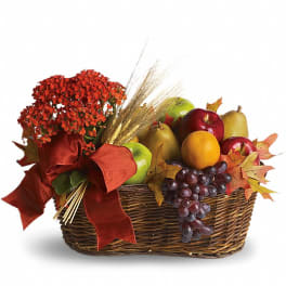 Wicker basket filled with assorted fruit, red flowers, wheat stalks, and a large red bow.