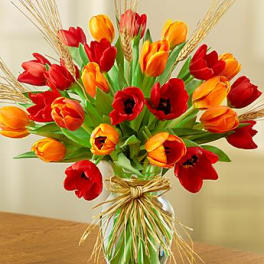 Red and orange tulips with wheat accents in a clear glass vase tied with raffia.