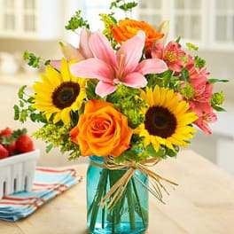 Bright bouquet of sunflowers, orange roses, and pink lilies in a blue mason jar vase with raffia bow