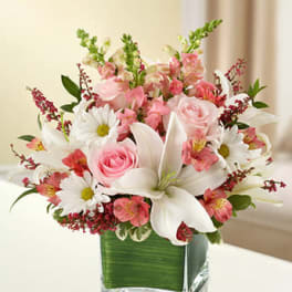 Pink roses, white lilies, and daisies in a square glass vase lined with green leaves