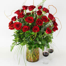 Red roses arranged in a glass vase with greenery and two wine glasses