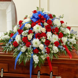 Patriotic red, white, and blue casket spray with roses, carnations, and mums topped with a large ribbon bow.