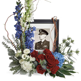 Red, white, and blue sympathy arrangement with roses, delphinium, and snapdragons around a framed military portrait