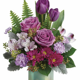 Arrangement of purple roses, tulips, and mixed pink and white flowers in an iridescent green glass vase