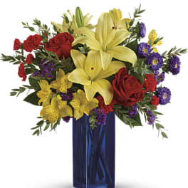 Tall blue vase with yellow lilies, red roses, red carnations, and purple and yellow accent flowers