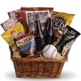 Wicker gift basket filled with assorted snack bags, two glass bottles, and a baseball.