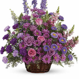 Basket arrangement of pink roses, purple carnations, and lavender daisies in a woven brown basket