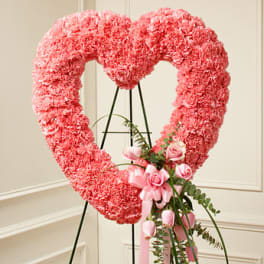 Standing pink carnation heart wreath with pink roses and ribbon on an easel.