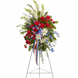 Tall standing spray of red carnations, white lilies, and blue flowers with patriotic ribbon on a metal easel.