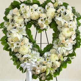 Heart-shaped wreath of white lilies, roses, and daisies on a stand with a white bow.