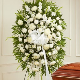 Tall standing spray of white flowers with large white bow on easel beside a wooden casket