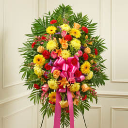 Tall standing spray of bright yellow, pink, orange, and red flowers with a large pink bow on an easel.
