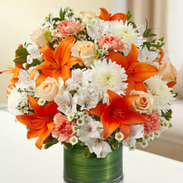 Round arrangement of orange lilies, white daisies, and peach roses in a green-wrapped glass vase
